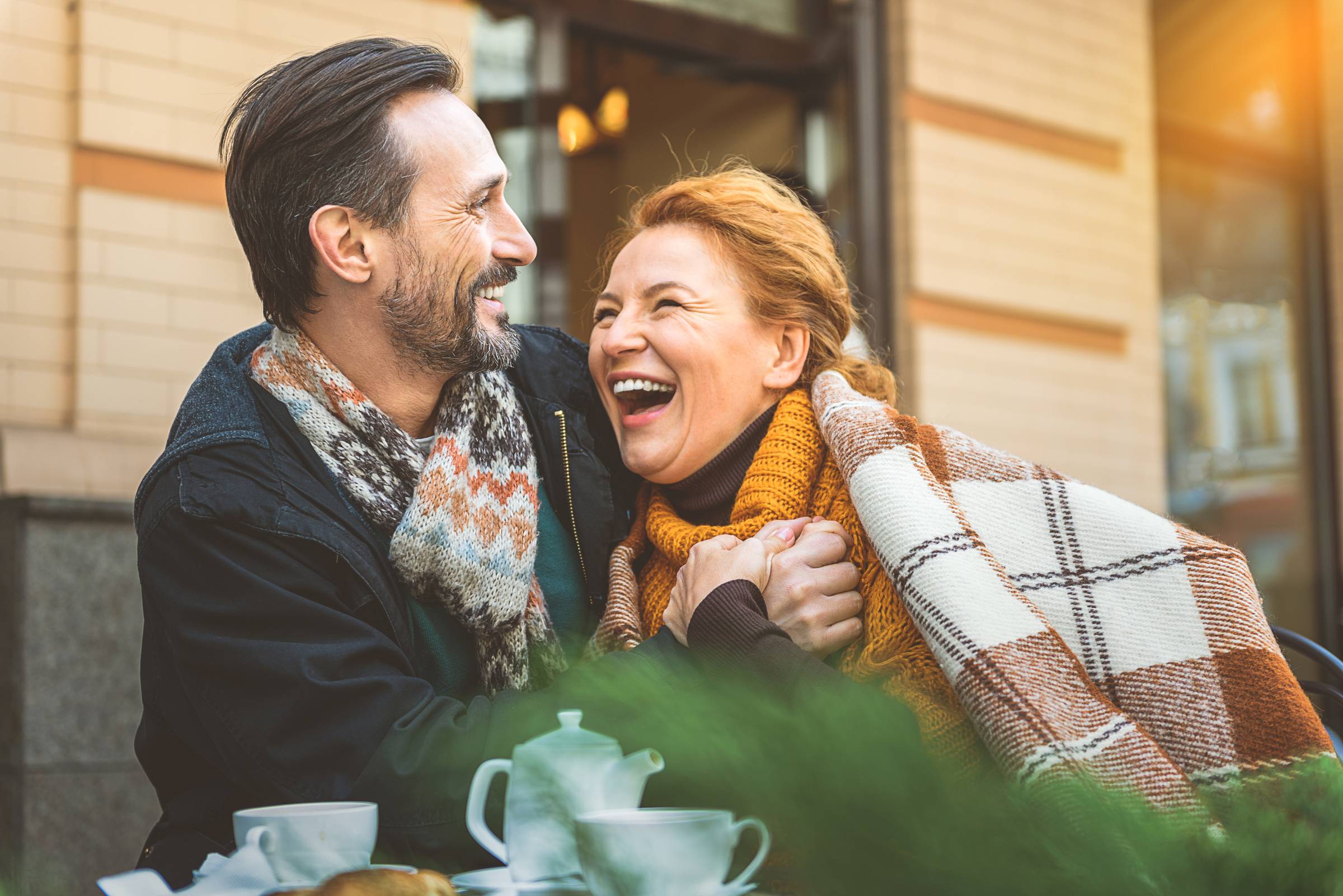 Royce Townhomes A happy couple in scarves laugh together at an outdoor café table with tea and pastries.