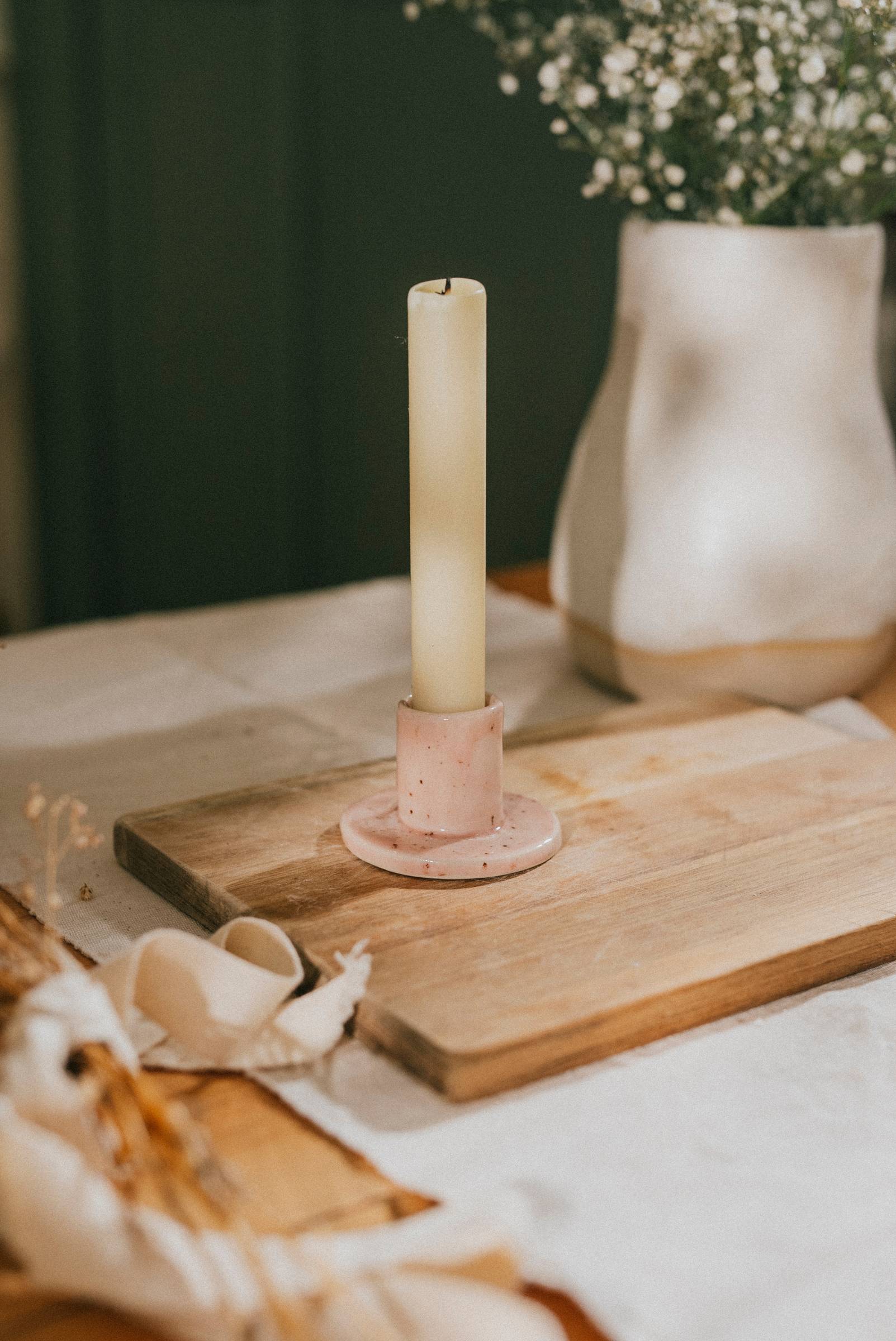 Royce Townhomes A tall white candle in a pink holder sits on a wooden board, with a white vase and table setting in the background.