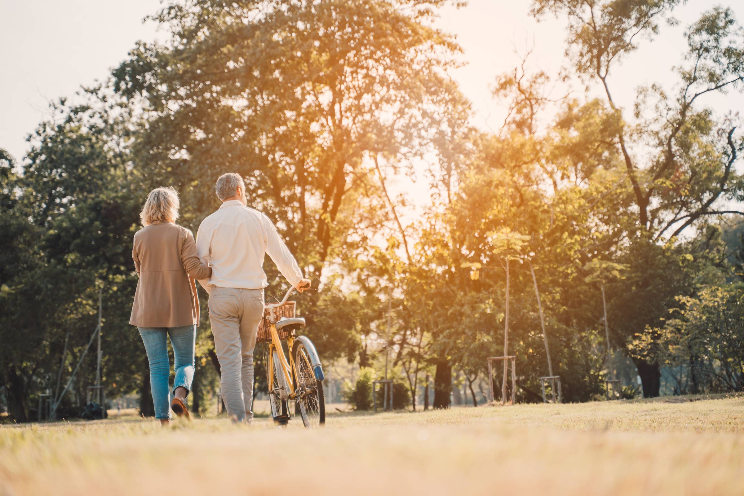 Royce Townhomes An older couple walks through a sunlit park, arm in arm, with a bicycle, surrounded by trees and grass.