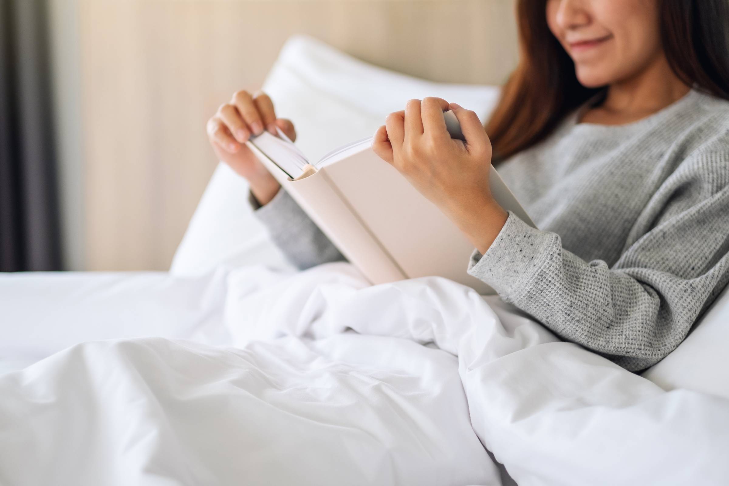 Royce Townhomes Woman in a gray sweater reading a book while sitting up in bed with white sheets and blankets.