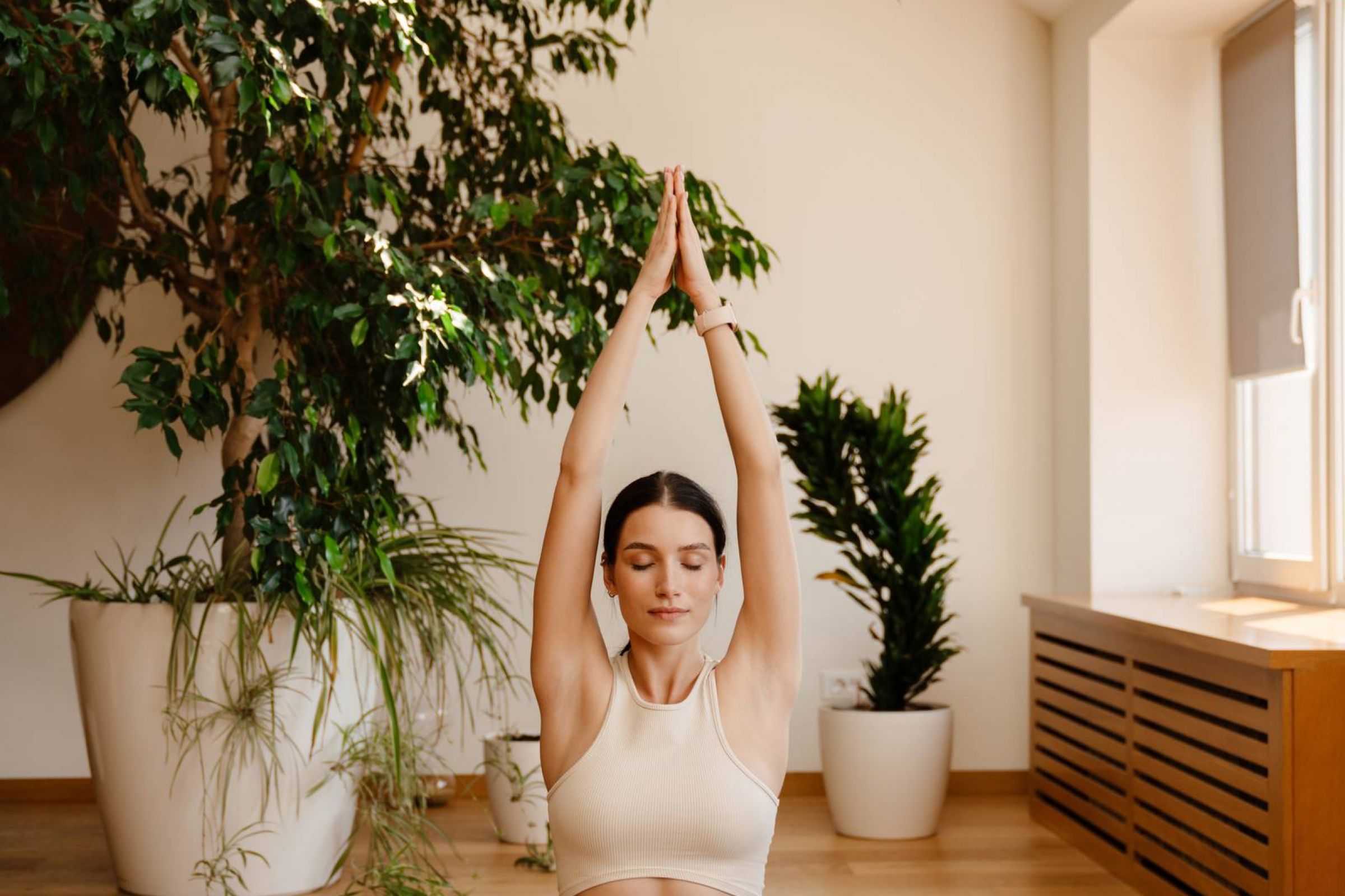 Royce Townhomes Woman in beige activewear practices yoga in lotus pose on a mat indoors, surrounded by green plants.