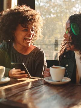 Two women laughing and talking at a café table with coffee cups, one holding a smartphone.