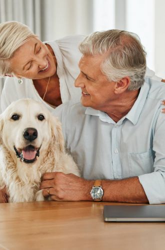 Royce Townhomes Smiling older couple embraces their happy golden retriever at a wooden table with a laptop and mug.