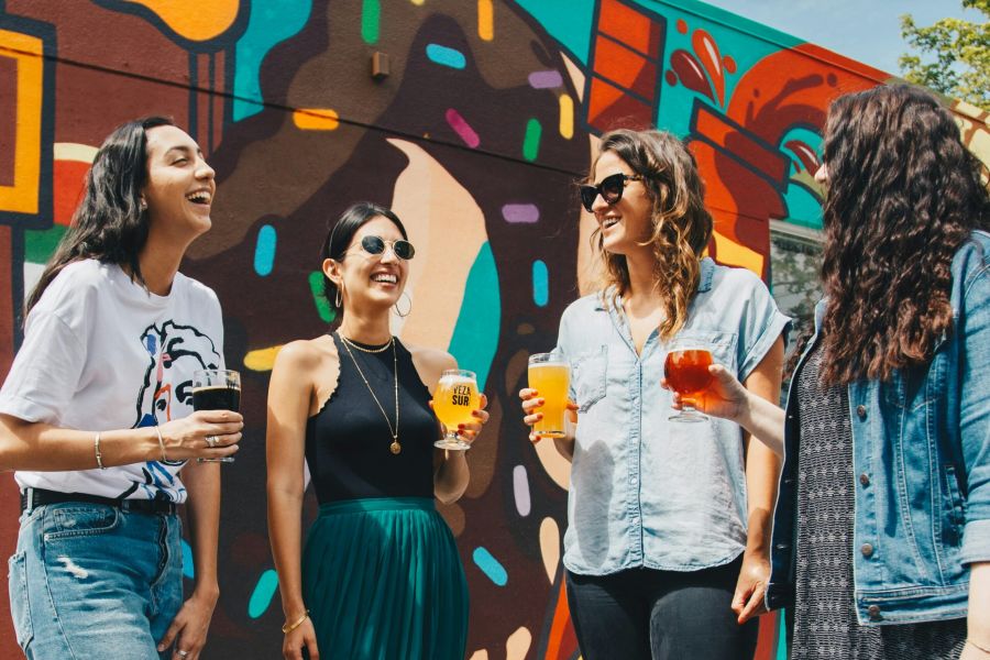 Royce Townhomes Four women laughing and holding drinks stand in front of a colorful mural on a sunny day.