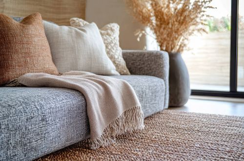 Royce Townhomes Gray sofa with neutral throw pillows and a beige blanket, beside a large vase of dried grass in a cozy room.