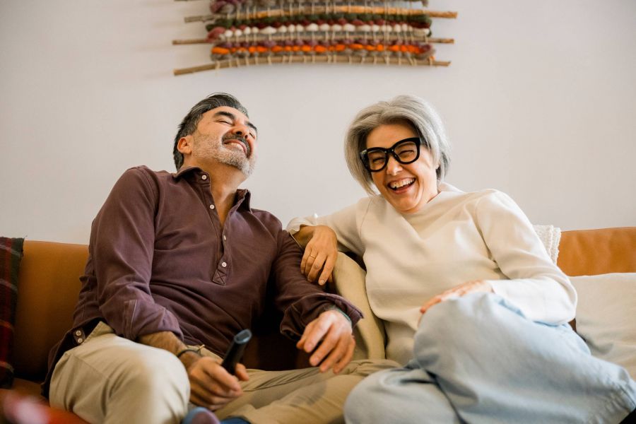 Royce Townhomes Two people sit on a couch laughing together, one holding a TV remote, with woven art hanging on the wall behind them.