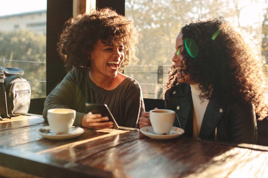 Two women laughing and talking at a café table with coffee cups, one holding a smartphone.