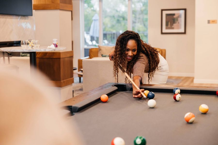 Royce Townhomes Woman smiling while aiming to take a shot at a pool table in a modern, well-lit living room.