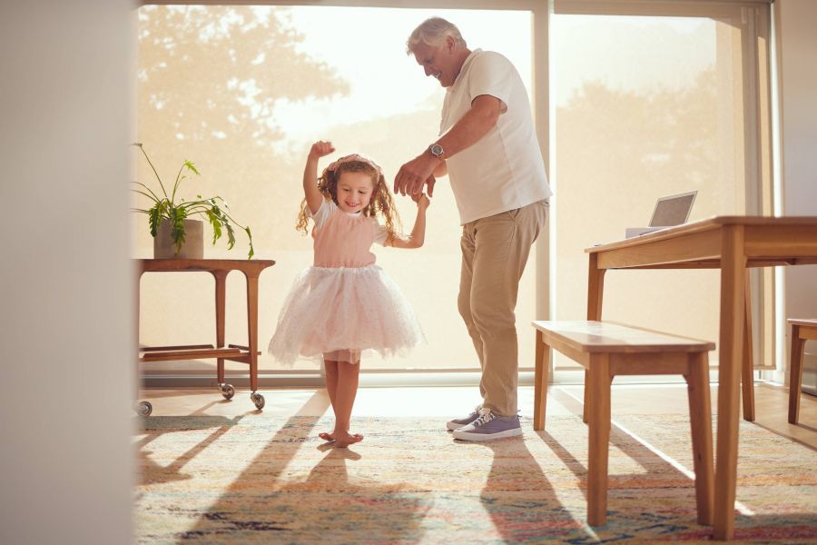 Royce Townhomes Older man and young girl dance together barefoot in a sunlit living room, smiling and holding hands near a table and plant.