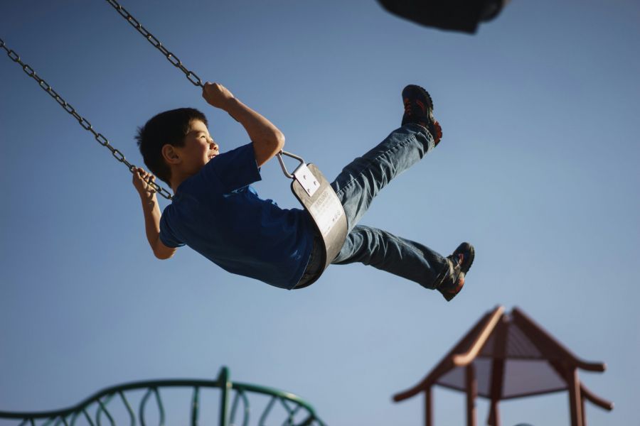 Royce Townhomes A young boy in jeans and a blue shirt swings high at a playground under a clear blue sky.