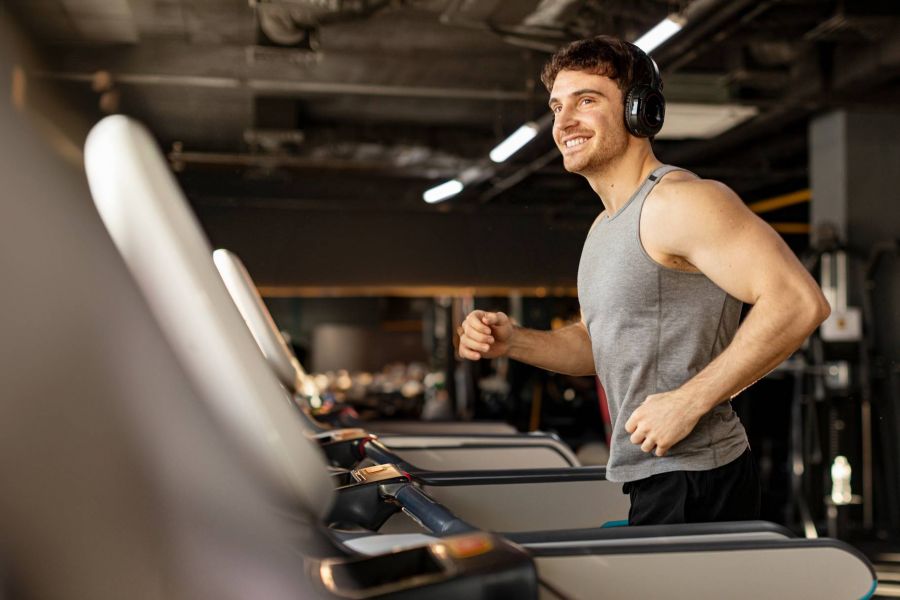 Royce Townhomes Smiling man wearing headphones runs on a treadmill in a gym, dressed in a gray tank top, with gym equipment in the background.