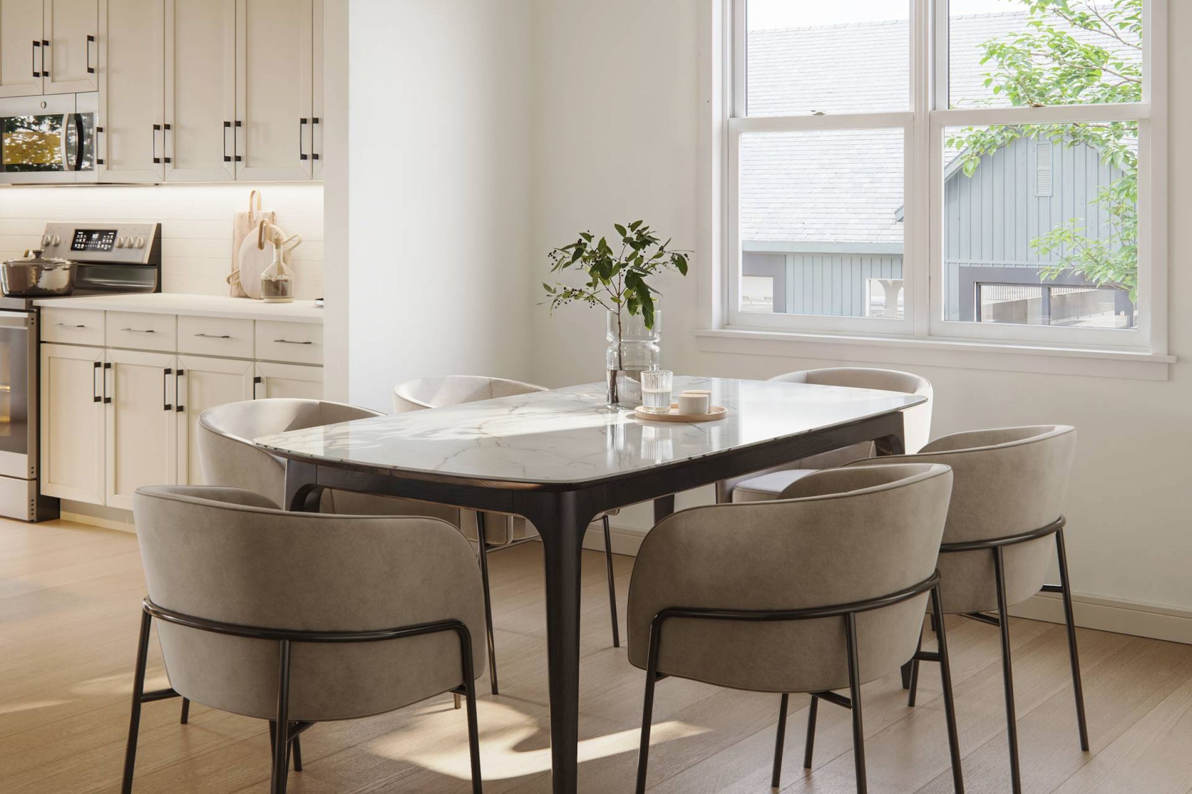 Royce Townhomes Modern dining area with a marble table, six chairs, and sunlight streaming through a window near a kitchen.